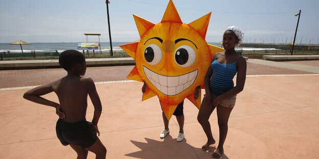Drei junge Menschen gruppieren sich an einer Strandpromenade um eine Art Sonnenlampion