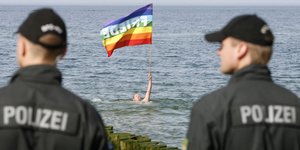 Ein Mann mit einer Regenbogen-Fahne schwimmt im Meer, zwei Polizisten beobachten ihn
