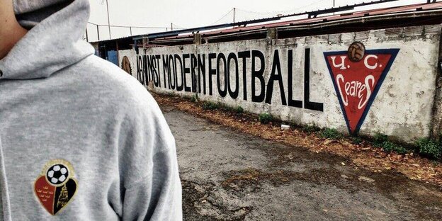 Im Vordergrund ein grauer Kapuzenpullover mit Vereinslogo, im Hintergrund steht auf einer Mauer "Against Modern Football"