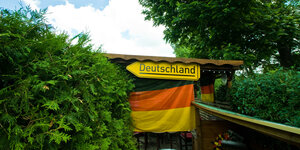 Ein gelbes Richtungsschild auf dem "Deutschland" steht hängt über einer Deutschlandflagge, daneben viel Grün