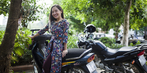 Eine vietnamesische Frau in einem geblümten Kleid lehnt an einme Motorroller