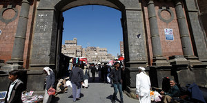 Blick durch ein Stadttor auf einen Markt in Sanaa, im Hintergrund die berühmten Lehmhäuser