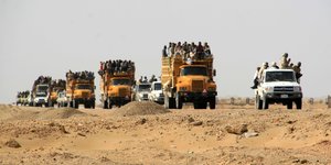 Eine Kolonne mit Fahrzeugen, auf denen sich viele Menschen drängen, fährt durch die Wüste