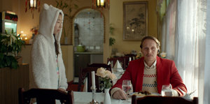 Ein Mann sitzt in einem Restaurant, neben ihm steht eine Frau in einem Kostüm