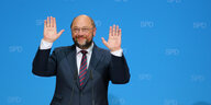 Martin Schulz vor blauer Wand, hebt beide Hände nach oben