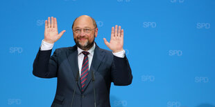 Martin Schulz vor blauer Wand, hebt beide Hände nach oben