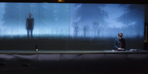 An Mann steht hinter einem DJ-Pult, dahinter eine verschwommen blaue Projektion, die Bäume und einen Menschen zeigt