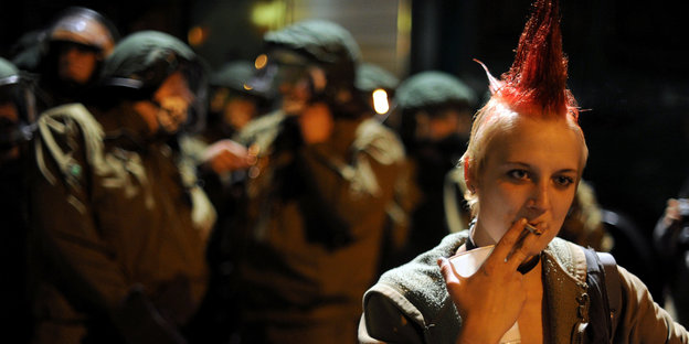 Junge Frau mit Punkfrisur raucht in der Nacht eine Zigarette, während dahinter Polizisten stehen