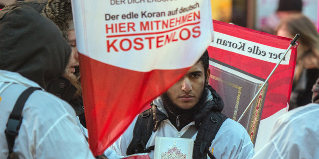 Männer in weißen Jacken, zwischen denen eine rot-weiße Fahne weht, auf der „Der edle Koran auf deutsch. Hier mitnehmen. Kostenlos“ steht.