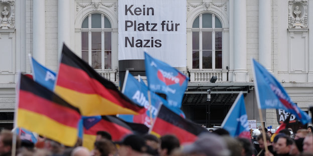 Menschen tragen Deutschland- und AfD-Fahnen, dahinter hängt ein Transparent an einem Gebäude, auf dem „Kein Platz für Nazis“ steht