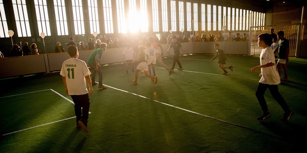 Ein Fußballfeld im Hangar des Tempelhofer Feldes in Berlin. Die Sonne scheint schräg zum Fenster rein. Kinder spielen im Gegenlicht.