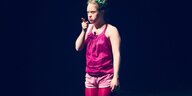 Eine Frau mit rosafarbener und pinker Kleidung steht auf einer Theaterbühne und zeigt mit einem Finger nach vorn. Sie hat das Down-Syndrom