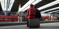 Eine junge Frau in roter Jacke und ein Mann sitzen auf einem Koffer und wartet auf den Zug