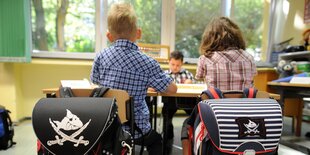 Zwei Grundschüler*innen von hinten, über den Stühlen hängen die Schulranzen, die Kinder brüten über einer Aufgabe