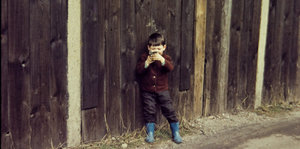 Ein kleiner Junge, der vor einem Bretterzaun neben einer Straße steht, richtet eine Pistole auf den/die Betrachter_in