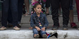 Ein Mädchen sitzt auf einem Bordstein, auf dem Erwachsene stehen und hält eine Kuba-Fahne in der Hand