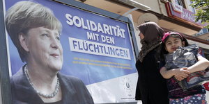 Auf eine Plakat ist Angela Merkel und der Satz „Solidarität mit Flüchtlingen“ zu sehen, davor steht eine Frau und ein Kind