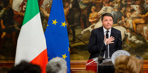 Ein Mann in Anzug und Krawatte steht vor einer Italien- und Europafahne