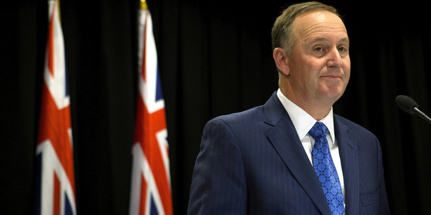 Ein Mann in Anzug und blauer Krawatte an einem Mikrofon, hinter ihm zwei neuseeländische Flaggen