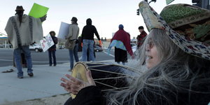Eine Frau mit Trommel sitzt auf dem Boden, mehrere Menschen mit Plakaten stehen in Reihen