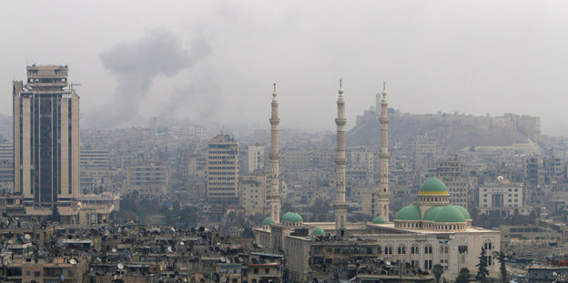 Eine Stadtansicht mit Minaretten und türkisfarbenen Kuppel, dahinter steigt Rauch auf