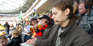 Auf der Tribüne eines Fußballstadions sitzen viele Menschen, im Vordergrund ein junger Mann, der in ein Headset spricht