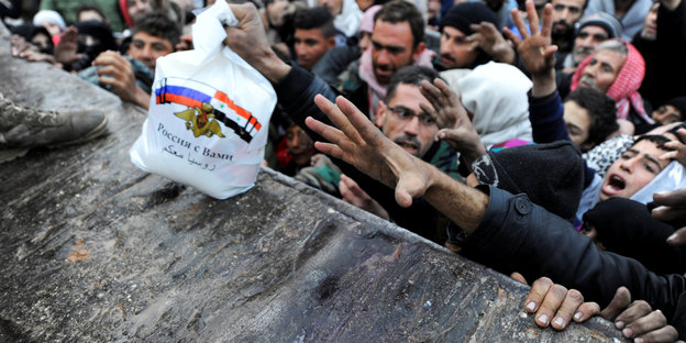 Eine Menschenmenge steht an einer Mauer, viele strecken die Hände aus, während ein kleines Paket mit kyrillischer Aufschrift herübergereicht wird