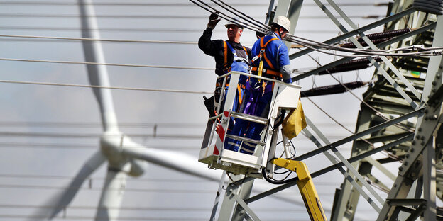 Vor der Kulisse eines Windrades arbeiten zwei Männer an Stromleitungen im Hochspannungsnetz