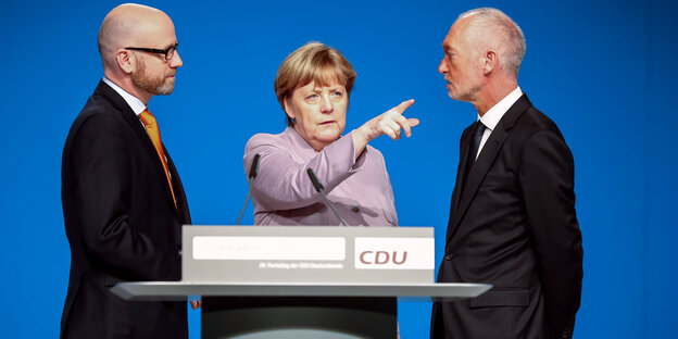 Angela Merkel hinter einem Rednerpult zeigt mit dem Finger in eine Richtung, rechts und links von ihr stehen Männer in Anzügen und sehen sie an