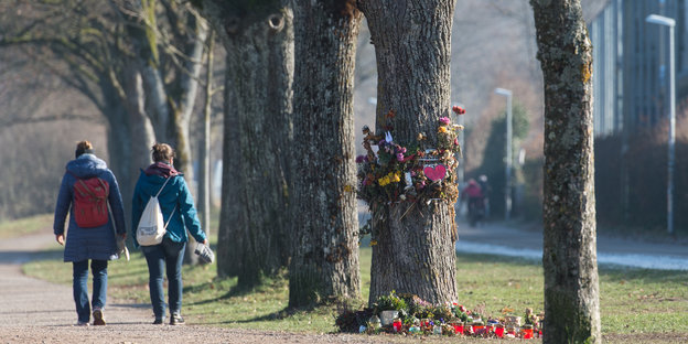 Spaziergänger gehen eine Allee entlang, an einem der Bäume am Wegesrand sind Blumen angebracht