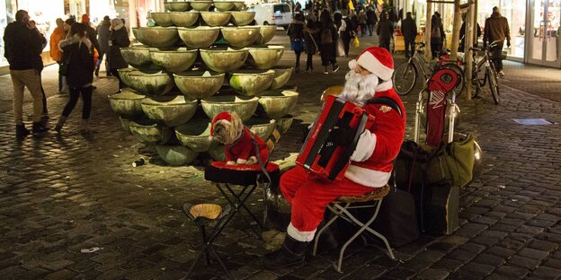 Ein Mann im Weihnachtsmannkostüm spielt Akkordeon auf einem Platz. Neben ihm sitzt ein Hund