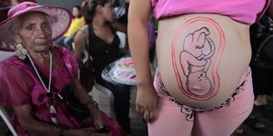 Auf den Bauch einer Schwangeren ist ein Baby gemalt, daneben sitzt eine alte Frau