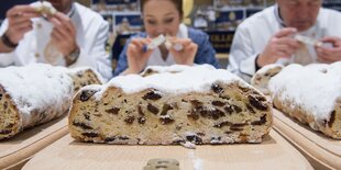 Ein angeschnittener Christstollen, im Hintergrund Menschen, die ihn probieren