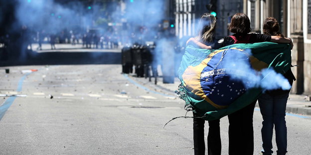 Demonstrantinnen mit Brasilien-Flagge stehen auf der Straße, im Hintergrund stehen Einsatztruppen