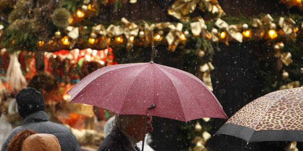 Menschen mit Regenschirmen laufen im Regen über den Wiehnachtsmarkt. Im Hintergrund ein Stand mit großer goldener Schleife