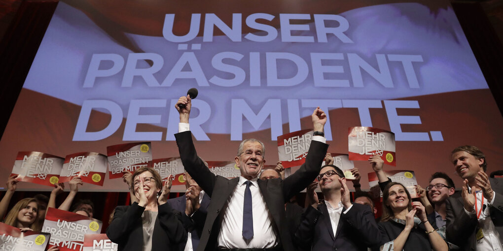 Van der Bellen hebt die Arme zur Siegerpose vor Parteifreunden und einer rot-weiß-roten Projektion an der Wand