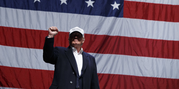 Donald Trump steht vor einer US-Flagge und ballt die Hand zur Faust