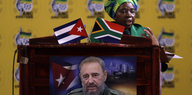 Eine Frau steht am Rednerpult, den eine südafrikanische und eine kubanische Flagge sowie ein Bild Fidel Castros schmücken