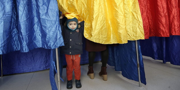 Ein kleiner Junge in einer Wahlkabine aus Stoff, die mit einer rumänischen Fahne verhangen ist
