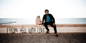 auf einer Mauer steht „FROM HELL WITH LOVE“ daneben sitzen ein kleines Mädchen und ein Mann