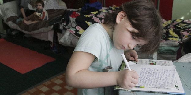 Ein Mädchen schreibt auf einem Blatt Papier. Im Hintergrund Betten