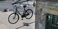 Ein Fahrrad hängt an einer Fassade. An einer Straßenecke sitzen zwei Personen.