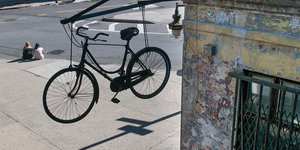 Ein Fahrrad hängt an einer Fassade. An einer Straßenecke sitzen zwei Personen.