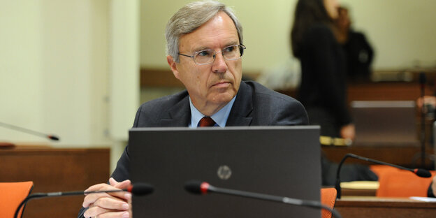 Ein Mann mit grauen Haaren sitzt im Anzug mit gefalteten Händen vor einem Laptop in einem Gerichtssaal