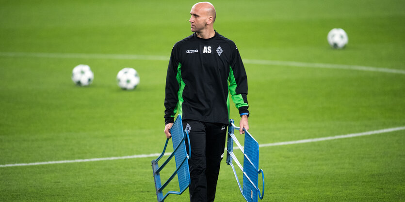 Ein glatzköpfiger Mann im Trainingsanzug geht über ein Fußballfeld