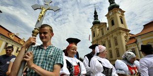 Ein junger Mann trägt ein Kreuz, an dem eine Jesusfigur hängt, dahinter läufen Frauen in Trachten