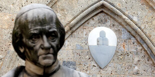 Der geschwärzte Männerkopf einer Statue vor einer Wand, an der ein Wappen hängt