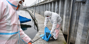 Zwei weiß vermummte Gestalten stehen auf einem Steg am Wasser packen etwas in einen blauen Abfallbeutel