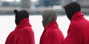 Drei Geflüchtete, die rote Decken über die Schultern gehängt haben vor einem Hafenbecken