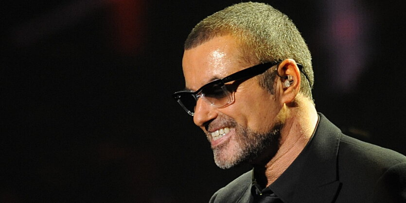 George Michael im schwarzen Anzug, mit schwarzer Sonnenbrille, vor schwarzem Hintergrund